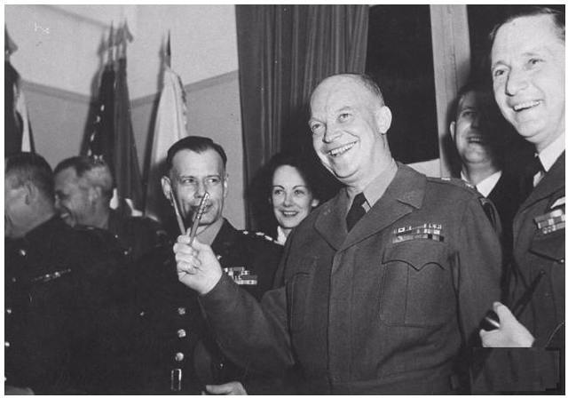 Un sonriente Dwight Eisenhower sosteniendo ds bolígrafos en forma de V de victoria. Detrás suya, Kay Summersby